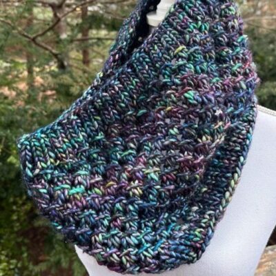A photo of a hand knitted Majestic Moraine textured cowl on a manikin outside.